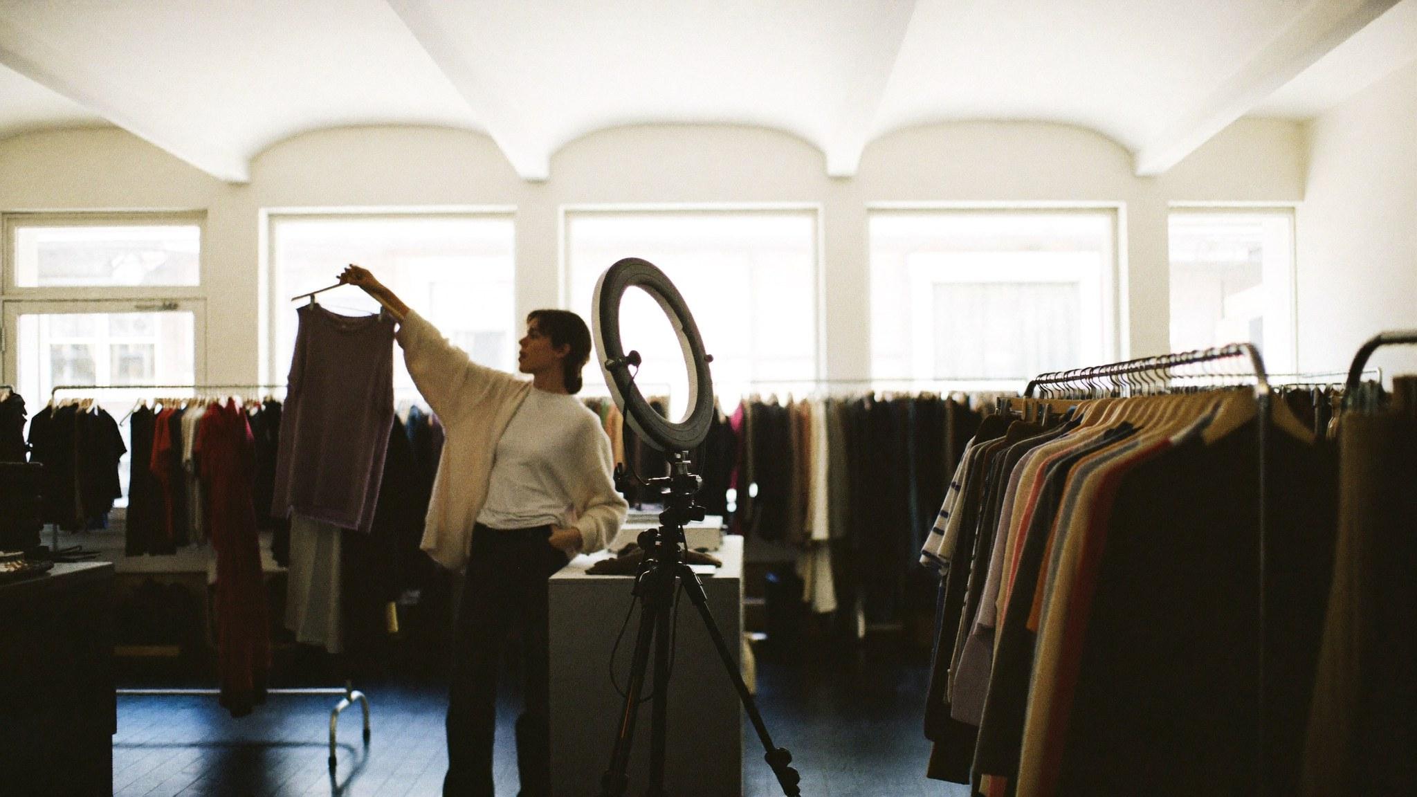 A live seller presenting a garment beside a lit ring light inside a bright boutique.