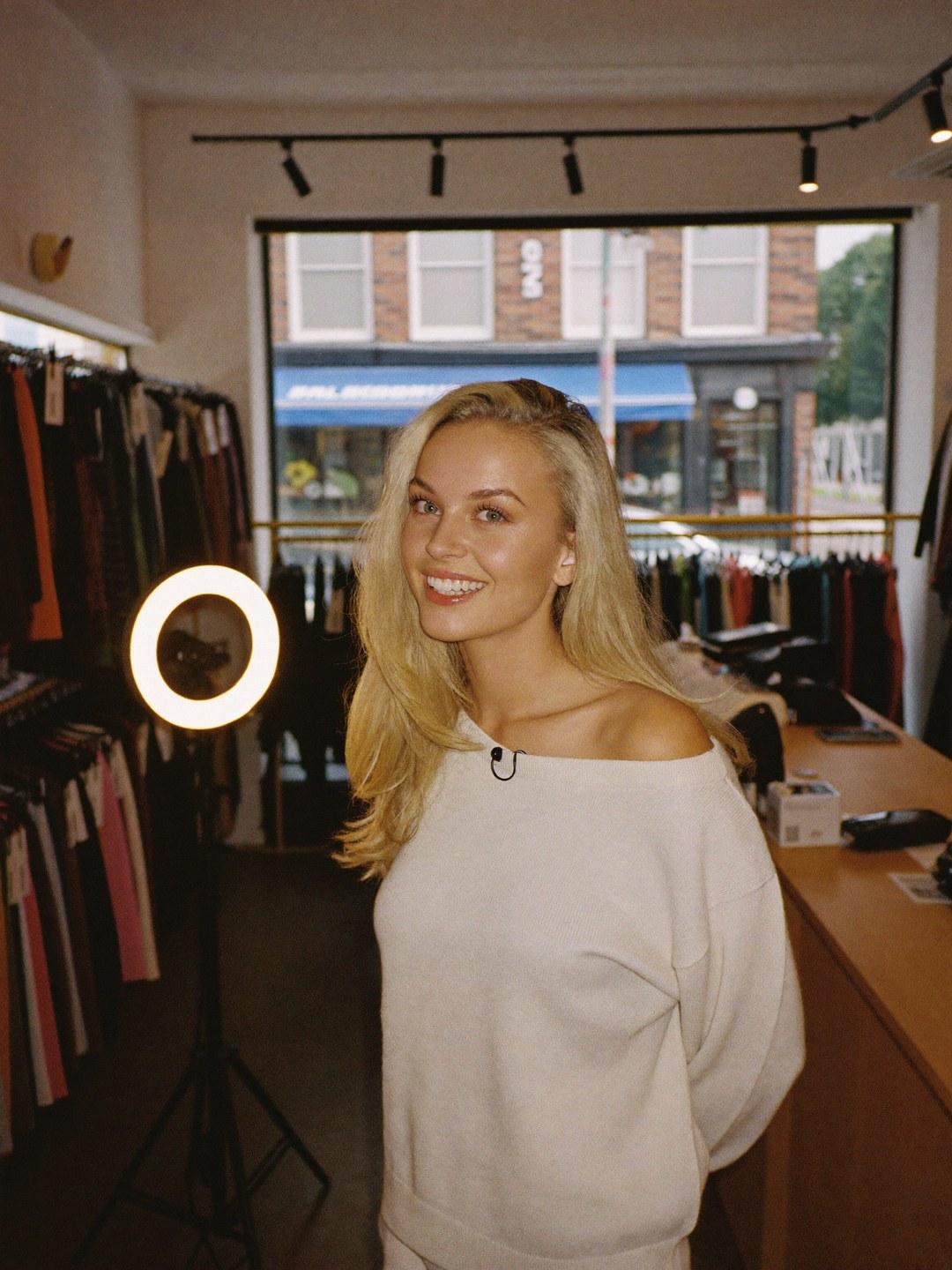 A host in a cream crewneck with a clip-on microphone, standing beside a ring light in a storefront.