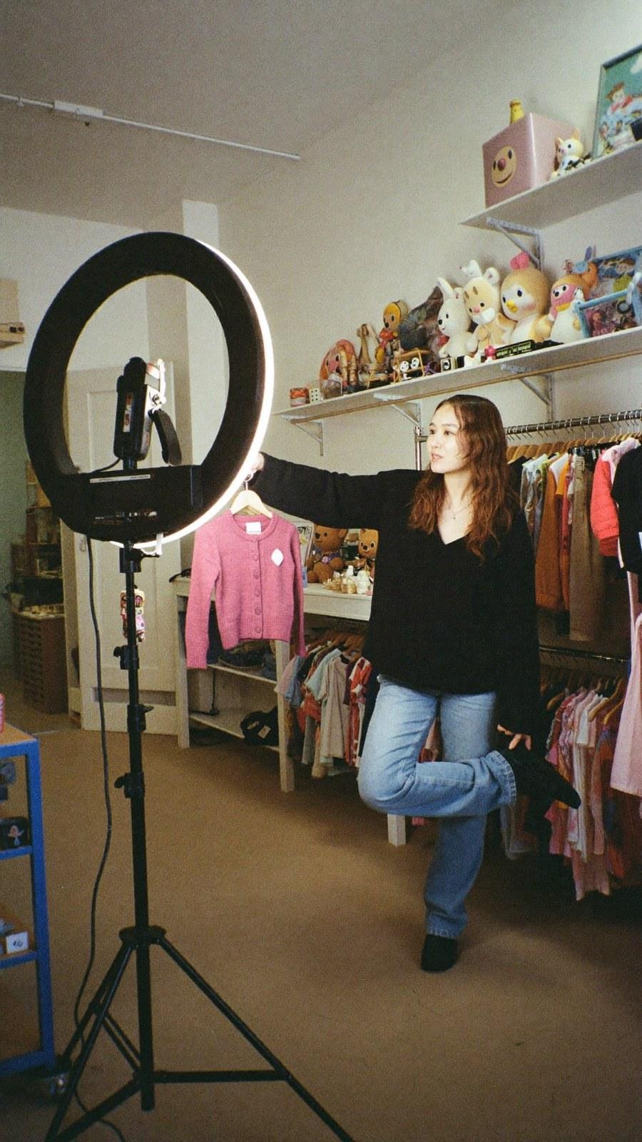A seller presenting a small pink cardigan beside a ring light in a children's boutique.